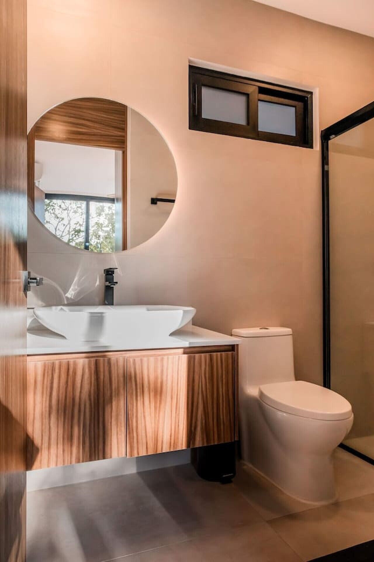 Ein modernes Badezimmer mit einem Holzkosmetikschrank, einem runden Spiegel, einem weißen Waschbecken und einer Toilette.