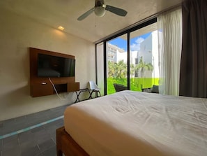 Ein Hotelzimmer mit Bett, Wandfernseher, Sessel und Balkon mit Blick auf Gebäude und Grünflächen.