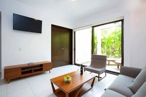 Ein modernes Wohnzimmer mit einem Flachbildfernseher, einem Couchtisch mit Früchten, einem grauen Sofa und einer großen Glasschiebetür, die in einen Garten führt.