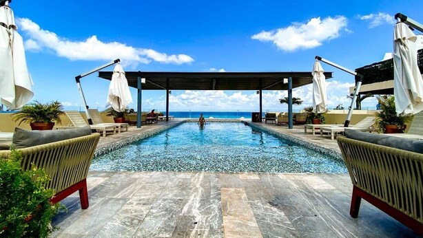 Ein Swimmingpool mit Liegestühlen und Sonnenschirmen, ein überdachter Bereich und Blick aufs Meer.