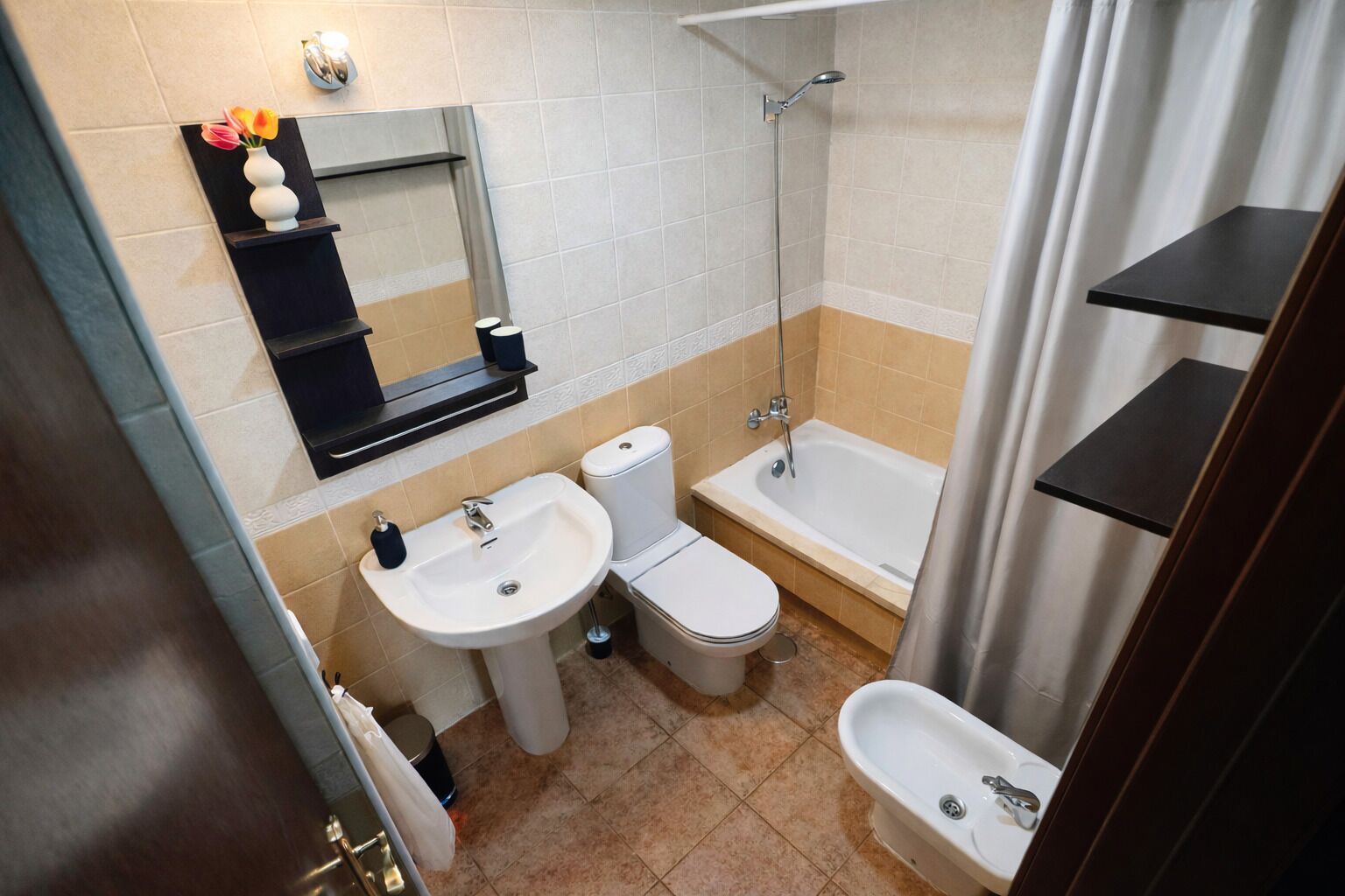 Une salle de bain équipée d’un lavabo, de toilettes, d’une baignoire et d’un bidet. On y trouve des étagères au-dessus du lavabo et un miroir au mur.