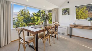 Un comedor con una mesa y sillas de madera, un florero con flores y vistas a árboles y montañas a través de amplios ventanales.