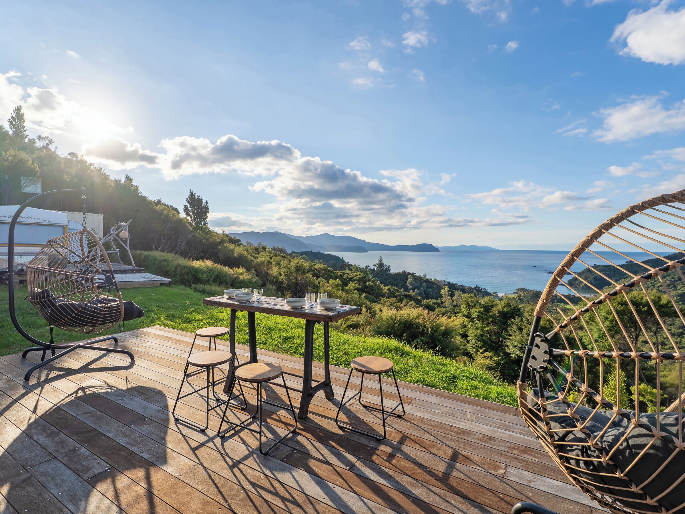 A wooden deck with a table and chairs, a hanging chair, and a view of the sea and mountains.