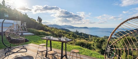 A wooden deck with a table and chairs, a hanging chair, and a view of the sea and mountains.