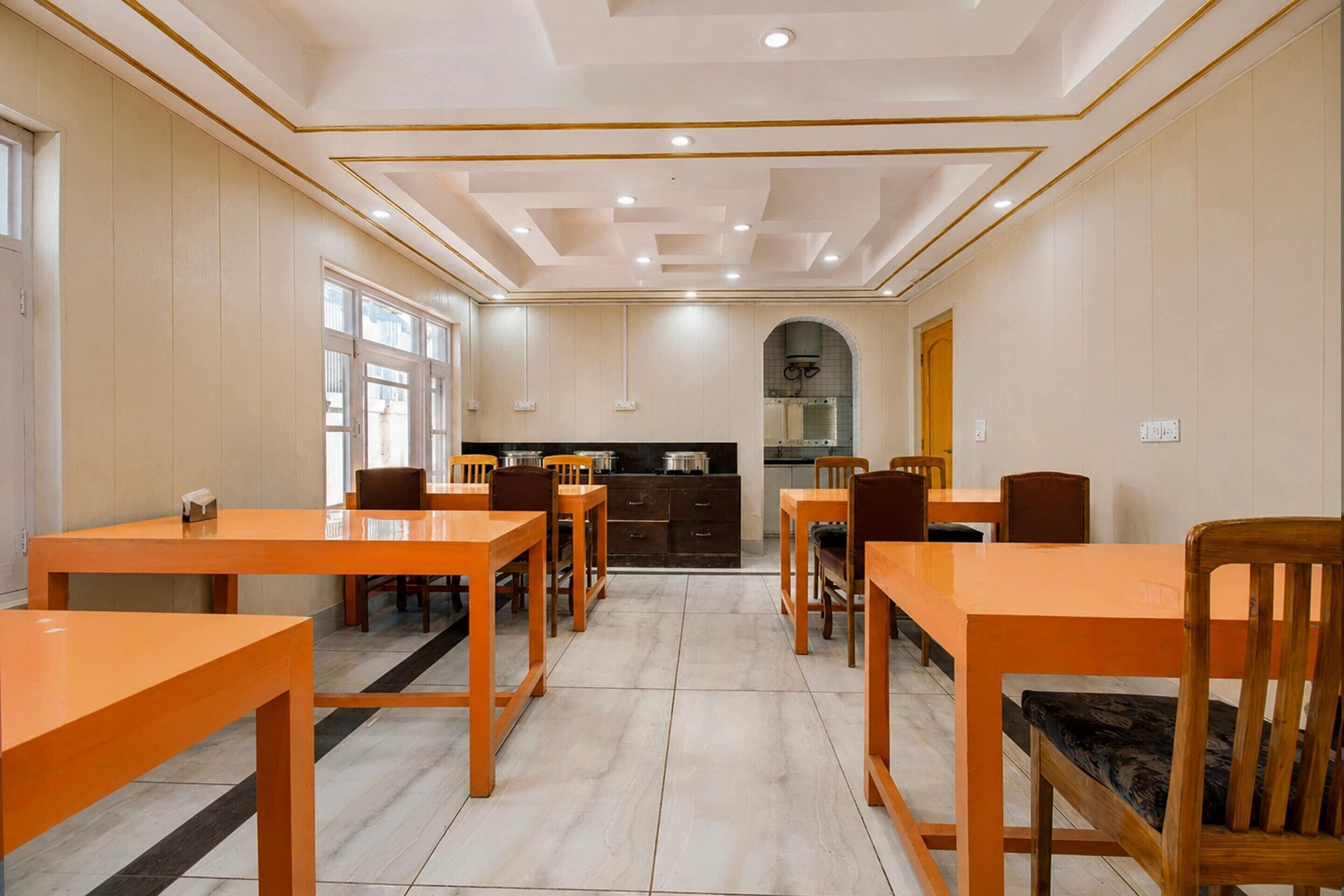 A dining area with wooden tables and chairs, a kitchen in the background, and a tiled floor.