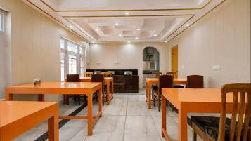A dining area with wooden tables and chairs, a kitchen in the background, and a tiled floor.