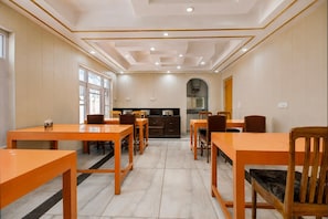 A dining area with wooden tables and chairs, a kitchen in the background, and a tiled floor. - Deluxe Pine Hills (Srinagar)