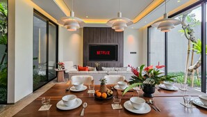 Ein modernes Wohnzimmer mit Netflix-Fernseher, bequemen Sitzgelegenheiten, einem Essbereich mit Tisch für vier Personen und großen Fenstern mit Blick auf Grünflächen.