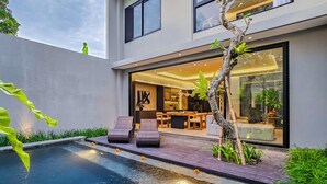 Ein modernes Haus mit Swimmingpool, Sitzgelegenheit im Freien und einer großen Glasdoor, die in einen hellen Innenraum führt.