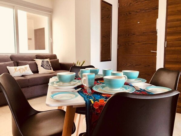 Un coin repas avec une table dressée pour quatre personnes, un canapé avec des coussins et, au fond, une porte en bois.