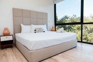 Ein modernes Schlafzimmer mit einem großen Bett, einem Nachttisch, zwei Nachttischlampen und Blick auf Bäume durch ein großes Fenster.