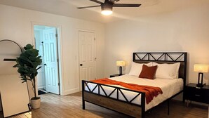 Ein Schlafzimmer mit einem Bett, einem Deckenventilator, einem Nachttisch, einer Lampe, einer Topfpflanze und einer Tür.