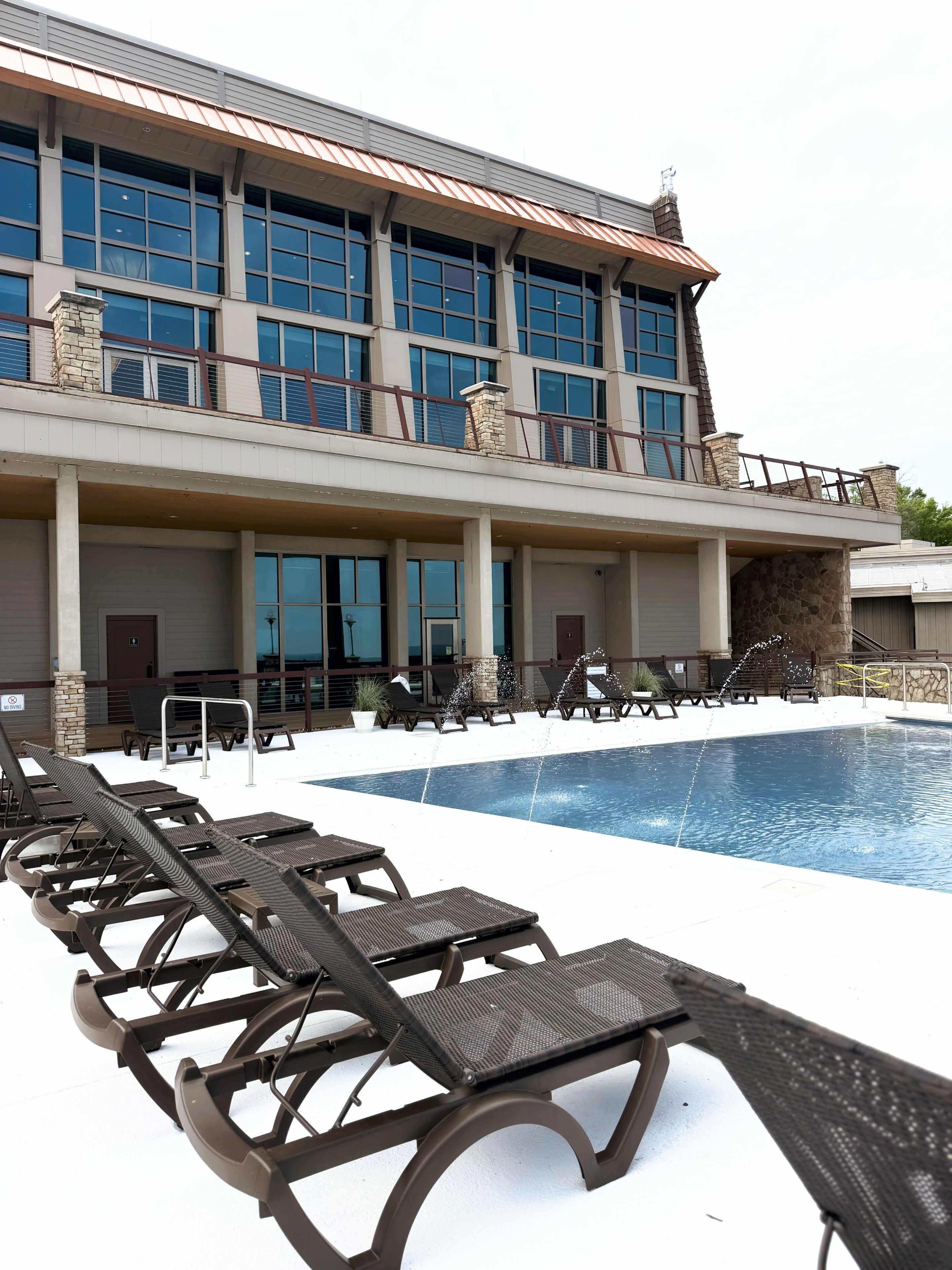 Un espace piscine avec des chaises longues et un bâtiment doté de nombreuses fenêtres et d’une façade en pierre.