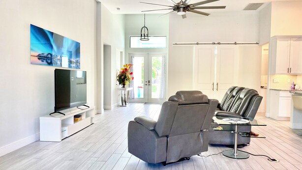 A modern living room with a flat-screen TV, a reclining chair, and a ceiling fan. - Gorgeous 3-bedroom house with Heated Pool and Spa (Cape Coral)