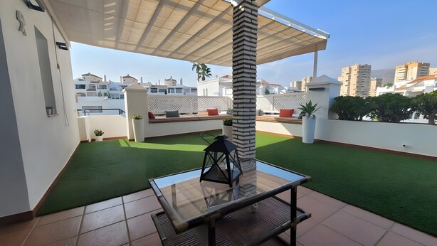 A rooftop terrace with a glass table, artificial grass, and a pergola.