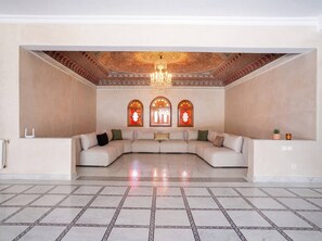 A spacious living area with a large L-shaped sofa, a patterned ceiling, and three arched windows with stained glass. - Villa Zitouna: Luxury Golf, Pool, and Garden in Amelkis + Free Airport Transfer  (Marrakech)