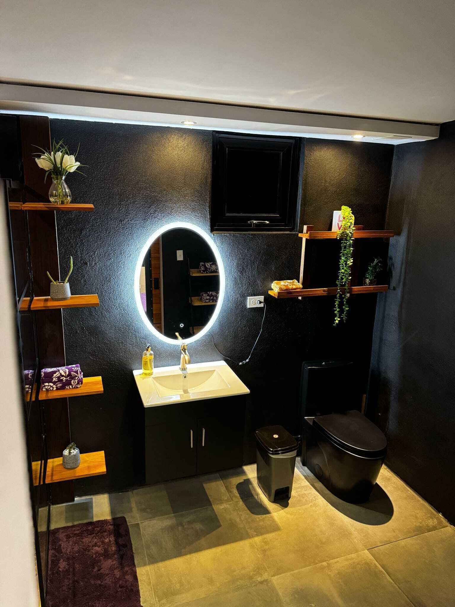 A modern bathroom with a round mirror, a sink, a toilet, and shelves with plants.