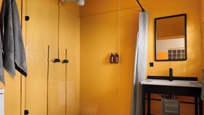 Een badkamer met een geel betegelde muur, een douche met gordijn, een wastafel met spiegel, en twee flessen zeep.