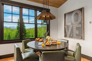 Uma sala de jantar com uma mesa redonda, cadeiras estofadas em verde, um candelabro e vista para montanhas através de grandes janelas.
