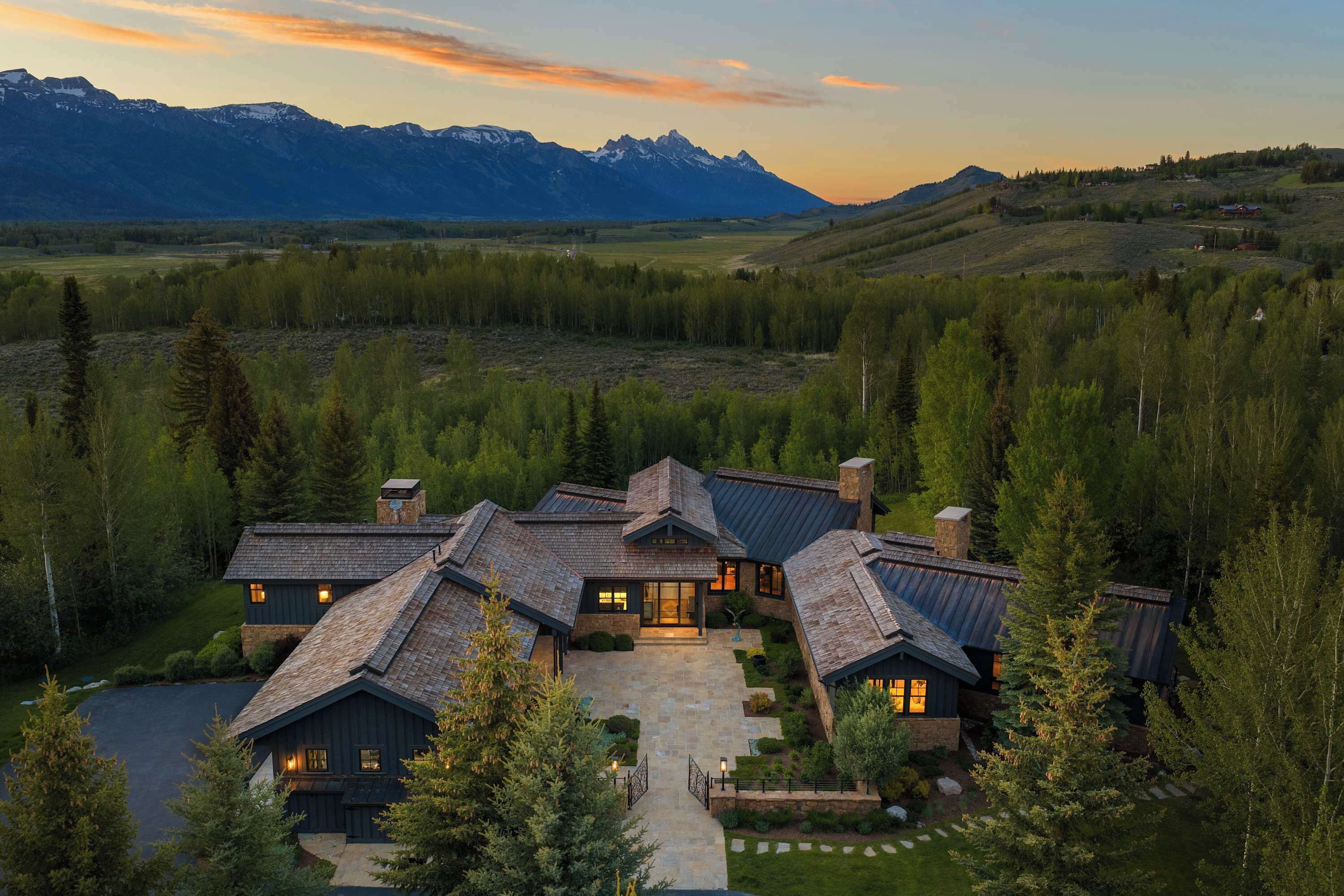 A multi-story house with a stone facade, surrounded by a forested landscape and mountains in the background.