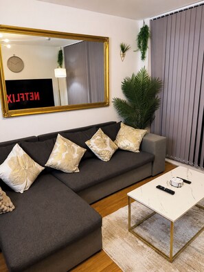 Ein modernes Wohnzimmer mit einem Ecksofa, einem großen Spiegel, einer Kübelpflanze und einem Couchtisch mit Fernbedienungen.