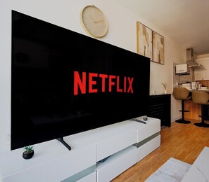 Ett modernt vardagsrum med en stor platt-TV som är monterad på en vit TV-bänk, en klocka på väggen och ett köksområde i bakgrunden.