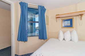 A hotel room with a bed, a window with blue curtains, a wall-mounted shelf with a picture, and a door.