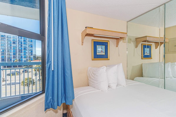 A hotel room with a large window, a bed with white linens, a blue curtain, and a framed picture on the wall.