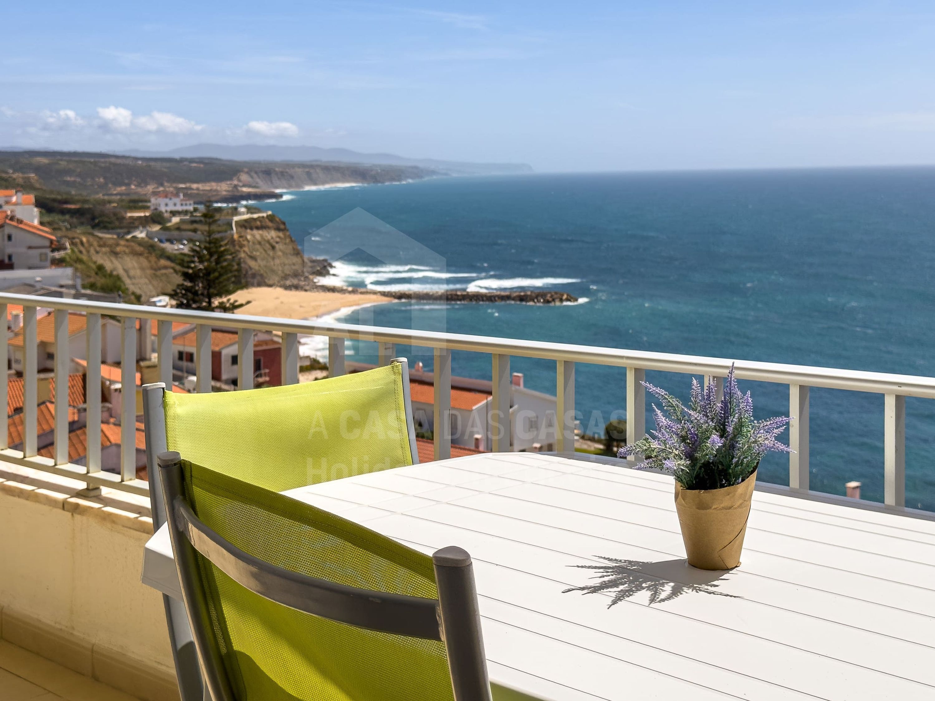 A balcony with a table and chair, overlooking a coastal view with a beach and ocean.