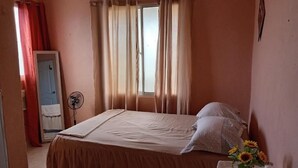 Une chambre à coucher avec un lit, des rideaux, un ventilateur, une porte et une petite table sur laquelle est posé un vase de fleurs.
