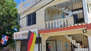 Un immeuble de deux étages doté d’un balcon, d’une entrée fermée par un portail et d’une enseigne ornée de drapeaux.