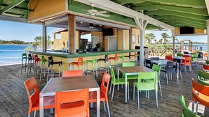 An outdoor dining area with wooden tables and chairs, a bar, and a view of the sea. - Bayside 1br BLOCK (Panama City Beach)