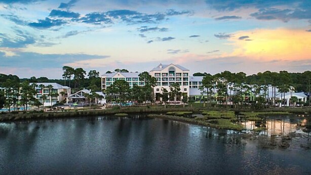 A waterfront hotel with multiple buildings, a lake, and a sunset sky. - Bayside 1br BLOCK (Panama City Beach)