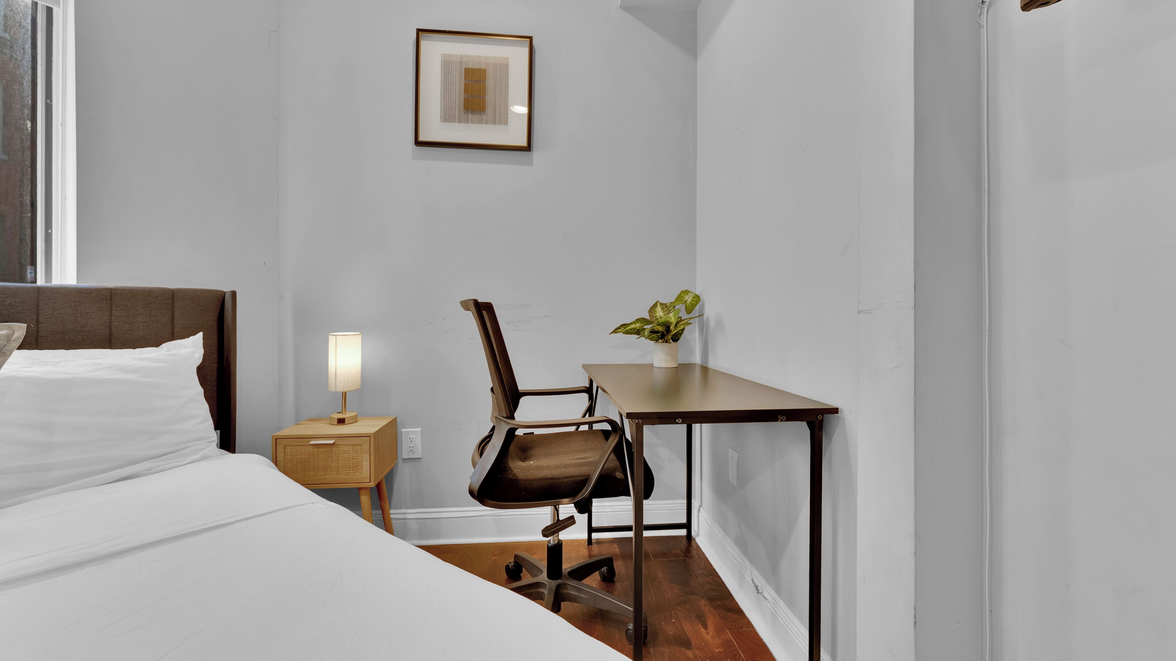 A hotel room with a bed, a desk, a chair, a lamp, and a framed picture on the wall.