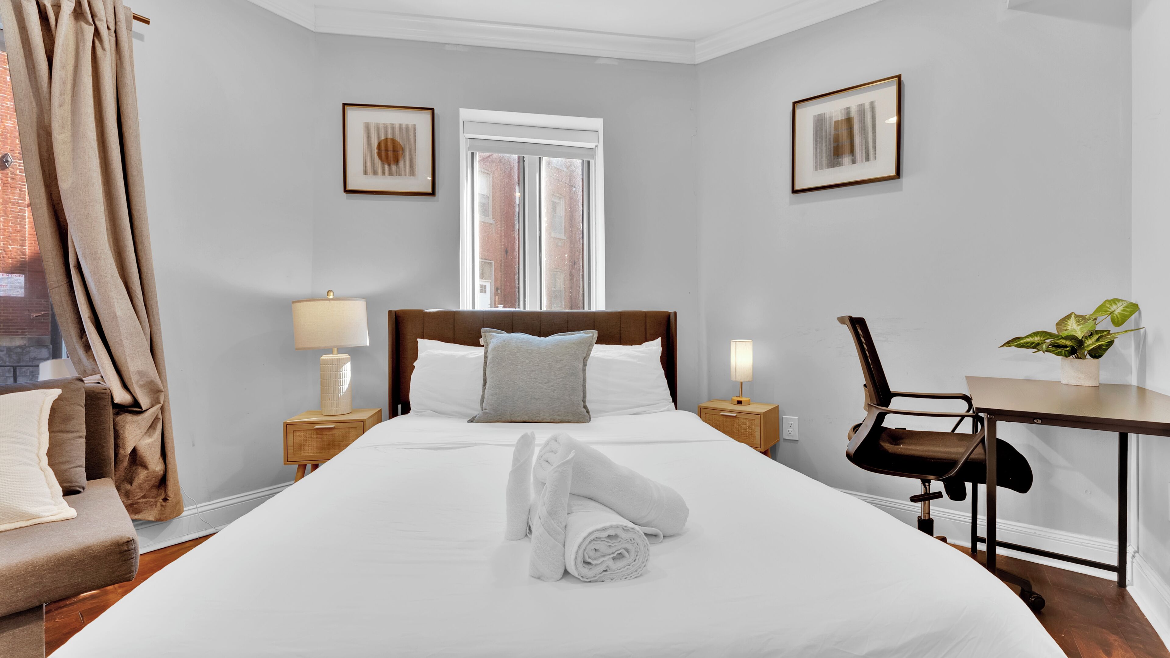 Un lit bien fait, avec du linge de lit blanc, une tête de lit en bois et un seul oreiller. Il y a deux tables de chevet avec des lampes, une chaise et une petite table sur laquelle est posée une plante.