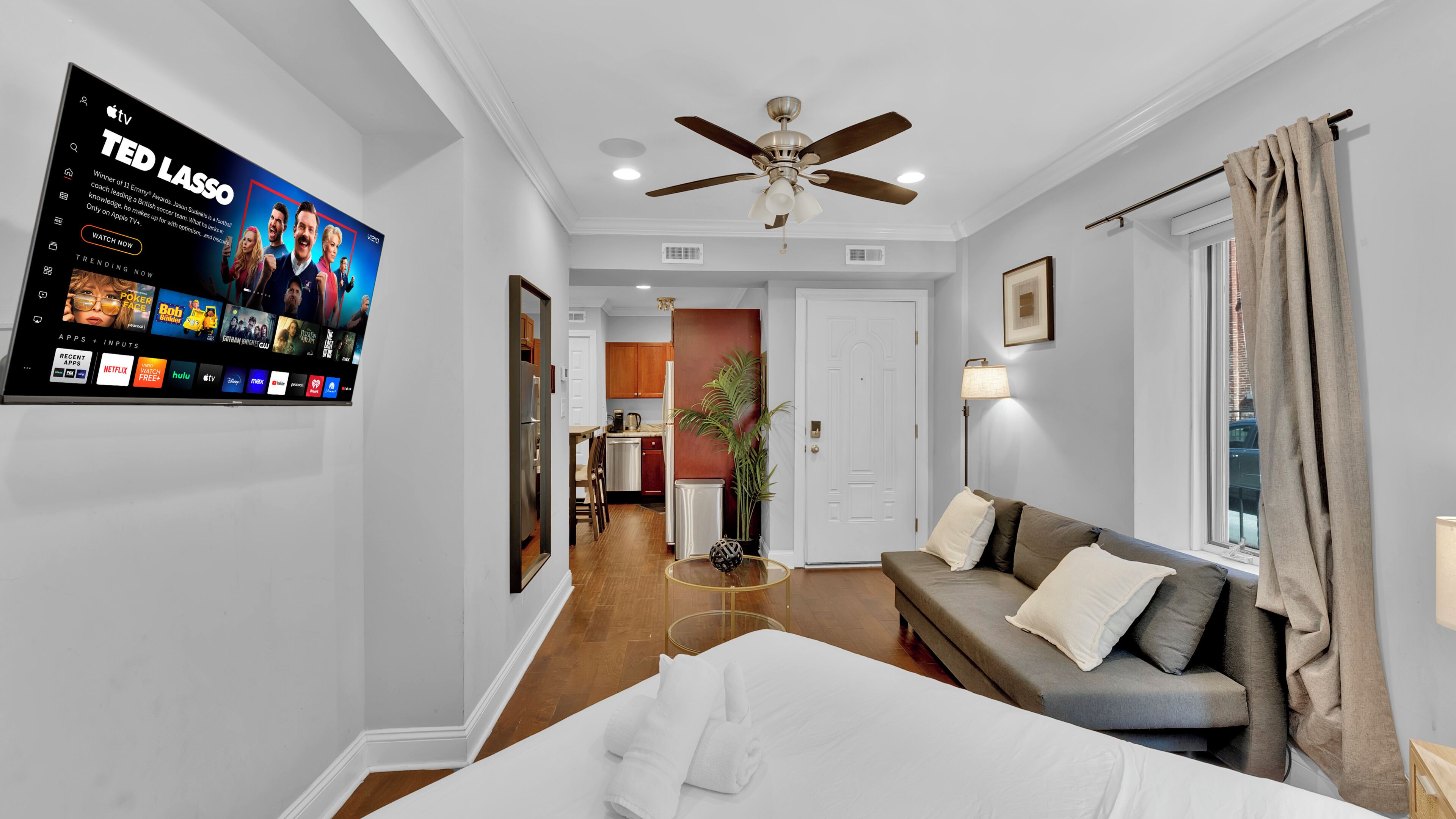 A hotel room with a bed, a sofa, a ceiling fan, a TV mounted on the wall, and a view of the kitchen and dining area.