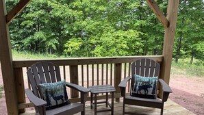 A wooden deck with two Adirondack chairs and a small table, surrounded by a wooden railing and lush green trees.