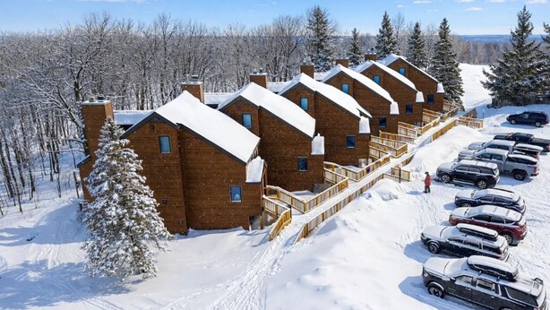 A row of wooden cabins with a snow-covered roof, a parking area with cars, and a forested background.