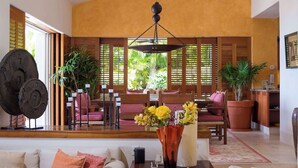 A spacious living area with a sofa, a coffee table, and a dining table set with chairs. There is a large window with wooden blinds, a hanging light fixture, and potted plants. - Four Seasons Villa Riva | 4BR Ocean-View Retreat w/ Infinity Pool & Golf Cart (Punta Mita)