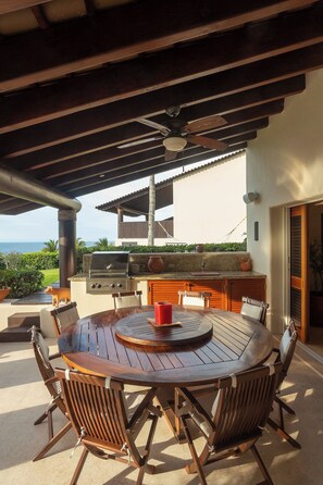 Eine Terrasse im Freien mit einem runden Holztisch, Stühlen, einem Deckenventilator und Blick auf den Ozean.