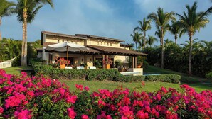A two-story building with a patio area, surrounded by lush greenery and palm trees. - Four Seasons Villa Riva | 4BR Ocean-View Retreat w/ Infinity Pool & Golf Cart (Punta Mita)
