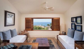 Ein Zimmer mit einem Bett, einer Couch, einem Couchtisch und Blick auf das Meer durch geöffnete Fensterläden.