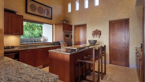 Cozinha com bancadas de granito, armários de madeira, ilha central com banquetas, janela com vista para o verde e porta com puxador decorativo.