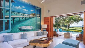 A spacious living area with a large painting of a tropical scene, a white sofa with blue cushions, a wooden coffee table, and a view of a golf course through large windows. - Luxury Golf-Front Villa: 6BR w/ Private Pool, Chef & Golf Cart (Punta Mita)