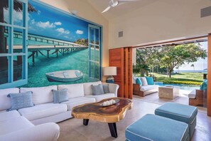 A spacious living area with a large painting of a tropical scene, a white sofa with blue cushions, a wooden coffee table, and a view of a golf course through large windows. - Luxury Golf-Front Villa: 6BR w/ Private Pool, Chef & Golf Cart (Punta Mita)