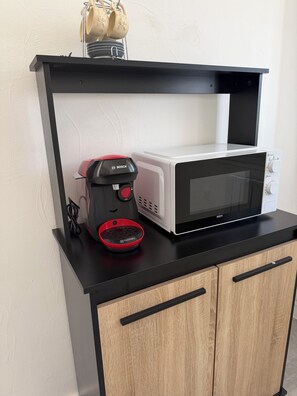 A kitchen with a microwave, a coffee maker, and a shelf with cups and a kettle.