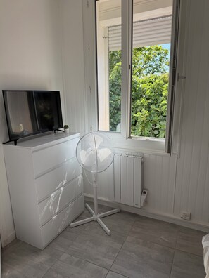 A room with a white radiator, a standing fan, a TV on a shelf, and a window with a view of greenery.