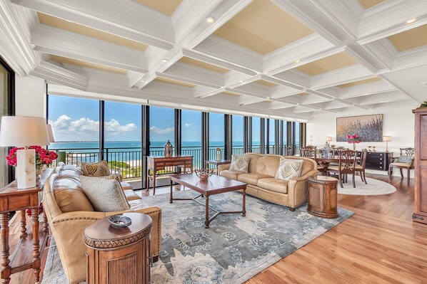 A spacious living area with a large window overlooking the sea, a sofa set, a coffee table, and a dining area with chairs and a table. - Mansions By The Sea 1201a - Premier (Treasure Island)