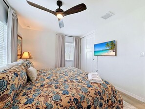 Ein Schlafzimmer mit einem Bett, einem Deckenventilator, einem Fernseher und einem Fenster mit Vorhängen.