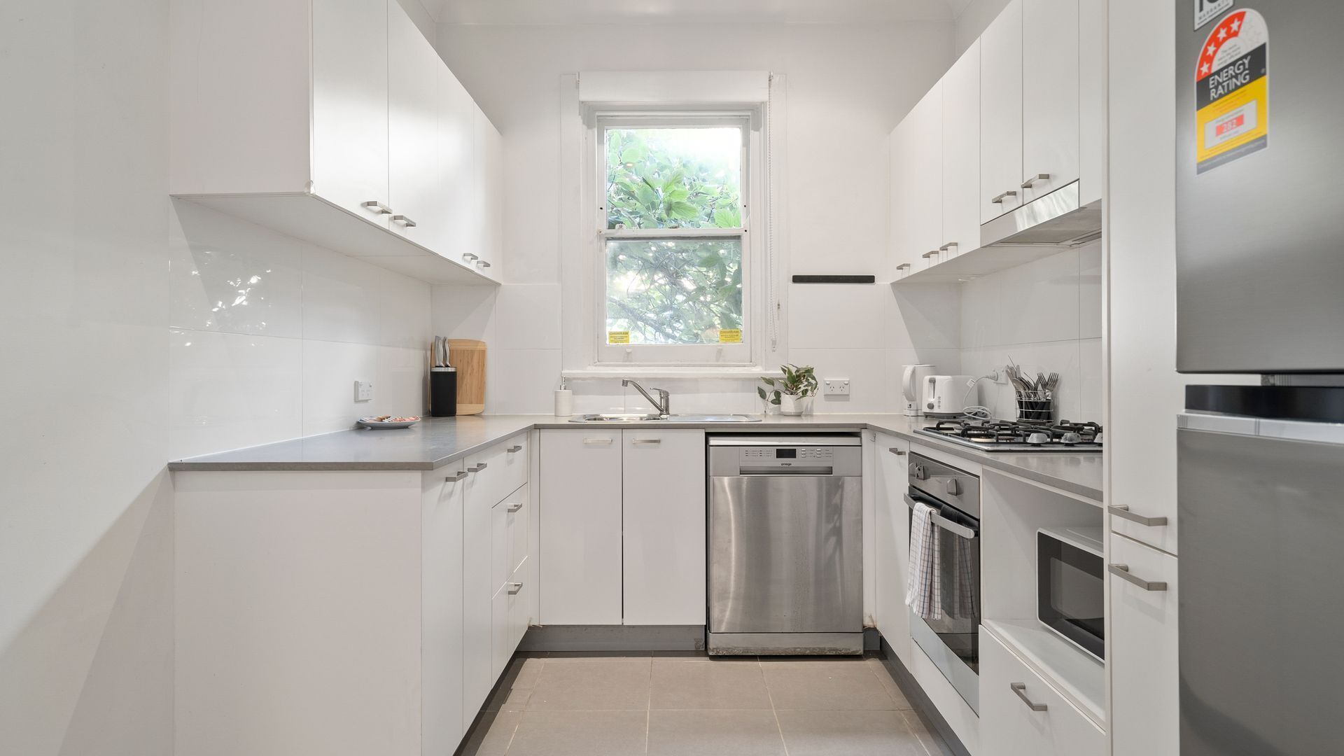 Eine moderne Küche mit weißen Schränken, einem Edelstahl-Geschirrspüler, einem Backofen und einem Fenster mit Blick auf Grünflächen.